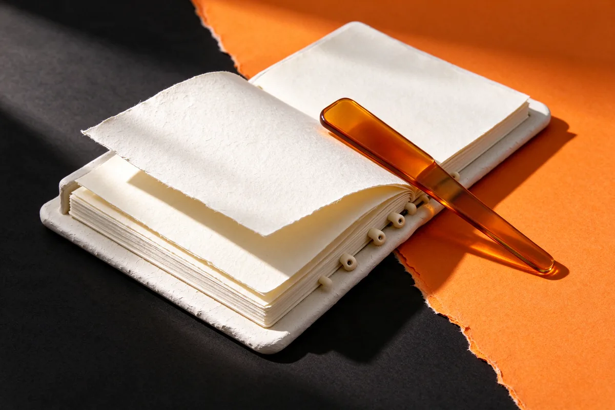An open white clay binder with loose pages and an amber glass bookmark on black and orange paper