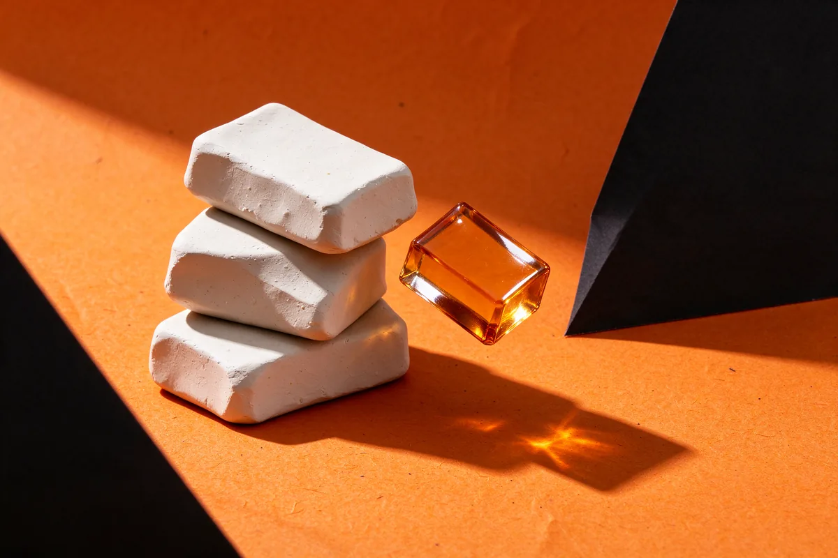 A tipped stack of white clay blocks with one amber glass block sliding off on orange and black paper